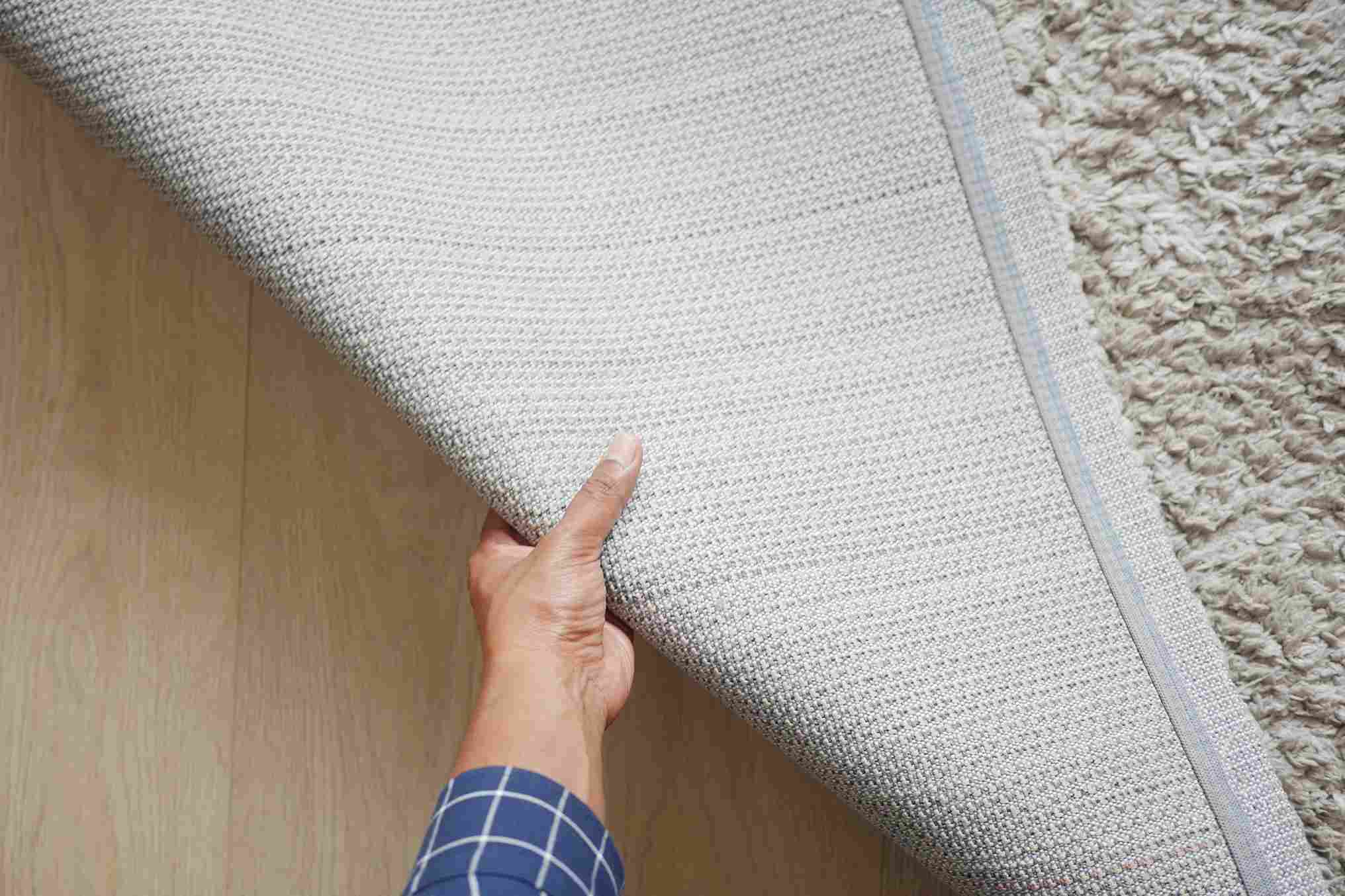 Ruka podiže ugao tepiha i prikazuje njegovu donju stranu i debljinu